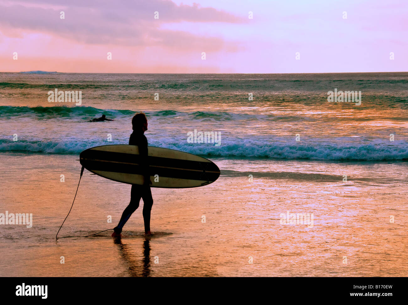 Un surfer carrying son conseil à marcher le long de la plage de Sennen à Cornwall au coucher du soleil. Banque D'Images