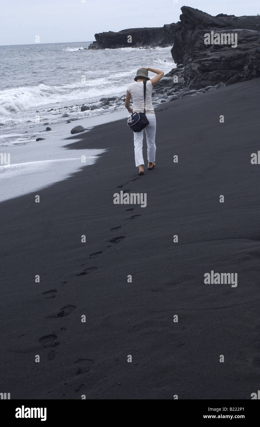 Une femme avec un sac photo marchant sur une plage de sable noir sur l'île d'Hawaii. Banque D'Images