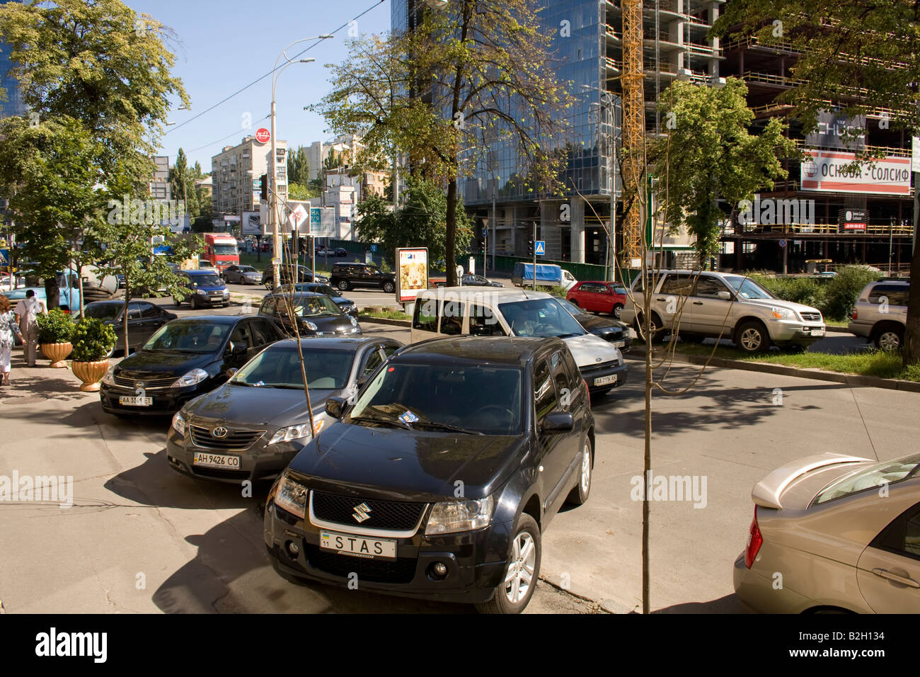 Rue animée à Kiev avec des voitures garées, la circulation, le chantier de construction et les arbres sous un ciel ensoleillé, mettant en valeur la vie urbaine et l'activité. Banque D'Images