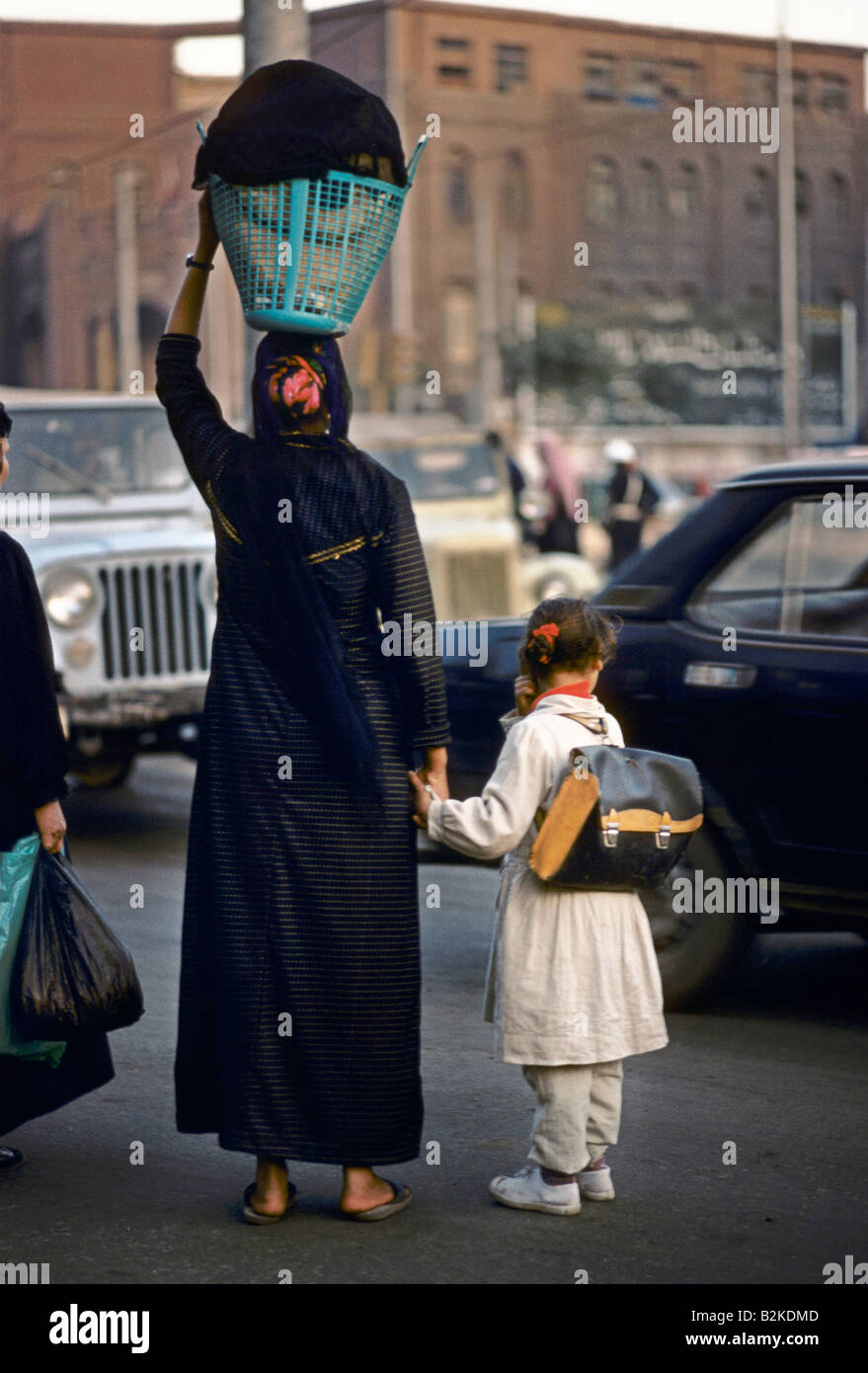 Scène de rue de la femme PORTANT SUR LA TÊTE PANIER HOLDING HAND D'ENFANT WAITIING À TRAVERSER LA ROUTE AVEC LE TRAFIC IMPORTANT LE CAIRE EGYPTE Banque D'Images