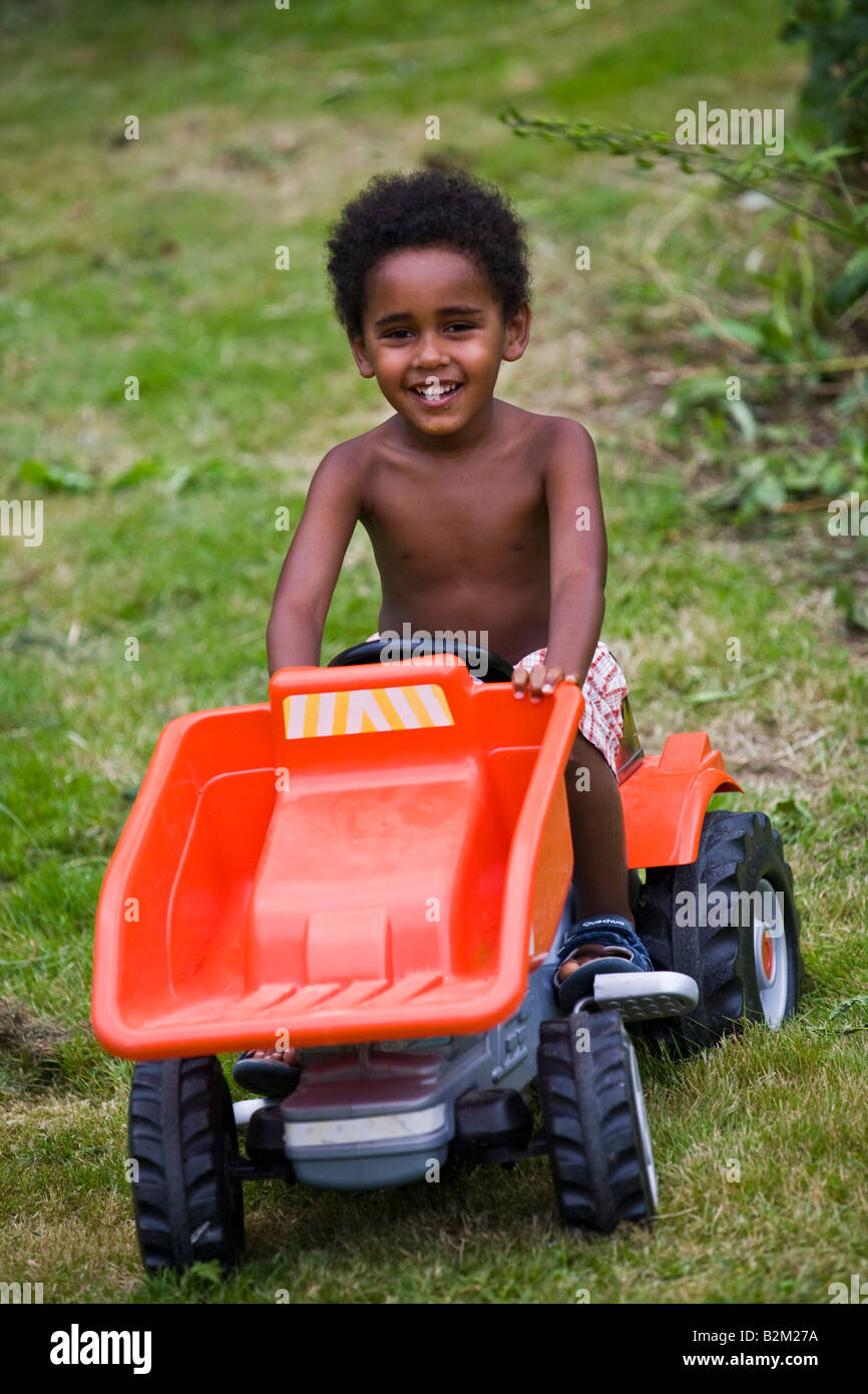 Un petit garçon noir au volant d'un jouet en plastique dans un jardin. Petit garçon de couleur conduisant un tracteur en plastique dans un jardin Banque D'Images