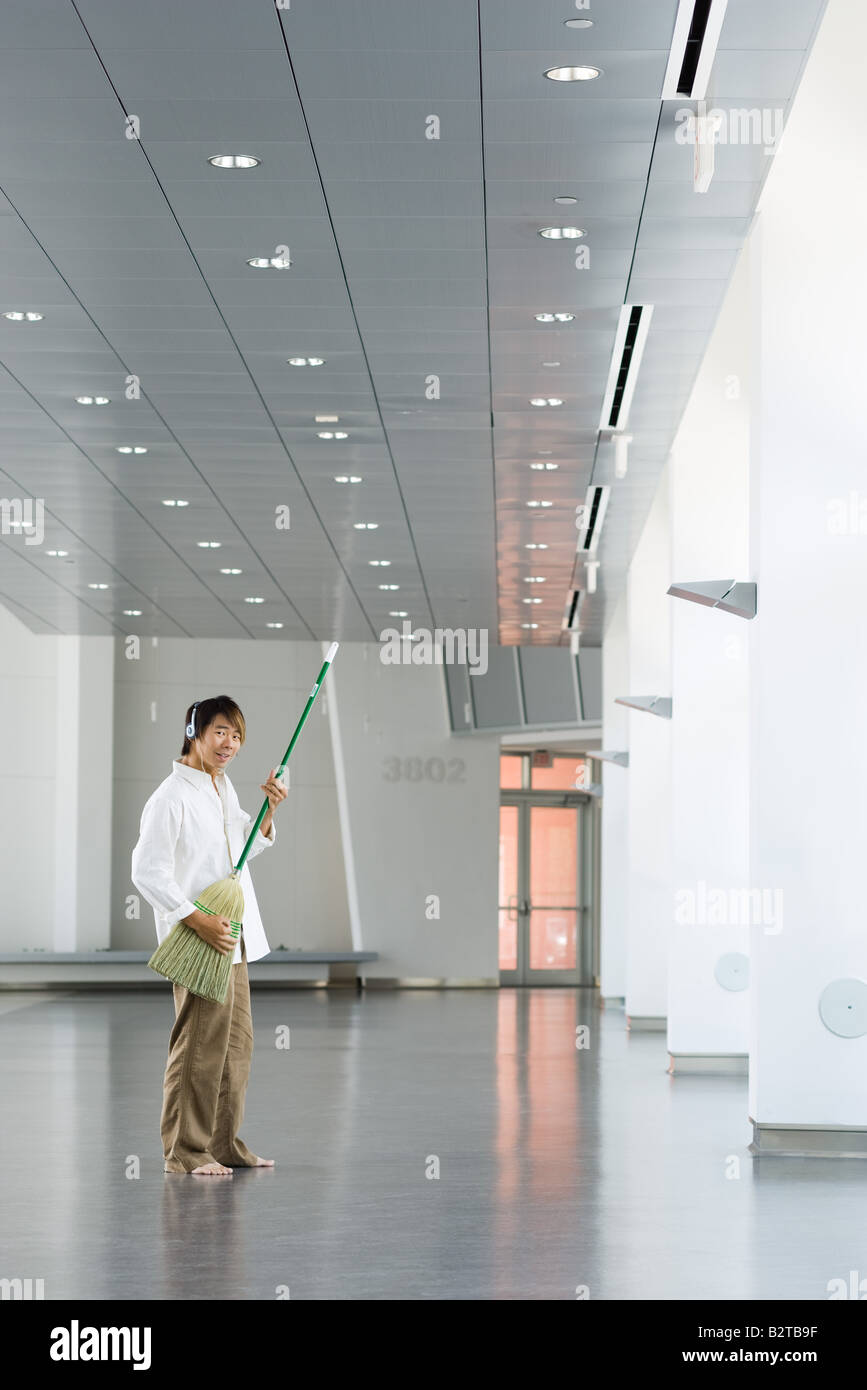 Homme debout dans le hall, playing air guitar avec balai, listening to headphones, smiling at camera Banque D'Images