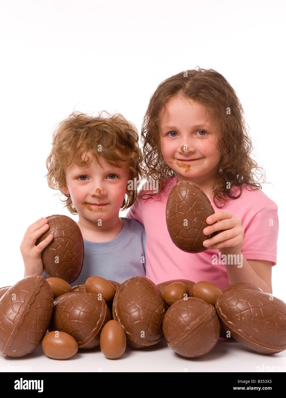 2 jeunes enfants avec du chocolat sur leur visage, chacun tenant un morceau d'oeuf de chocolat, avec beaucoup plus d'oeufs de Pâques au premier plan. Arrière-plan blanc Banque D'Images