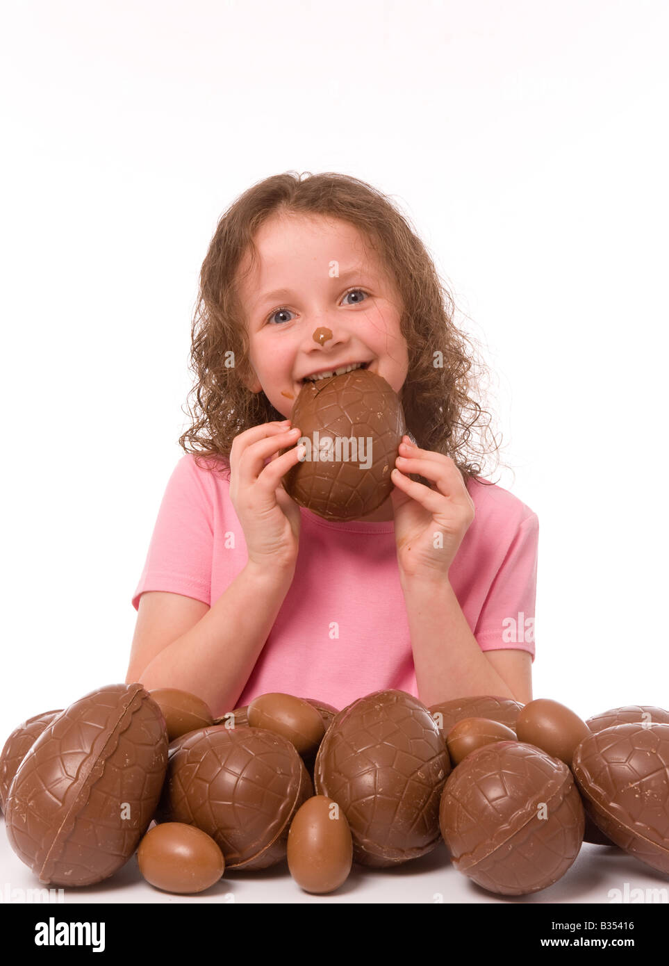 Jeune enfant portant un t-shirt rose avec du chocolat sur le visage, avec une grosse pile d'oeufs de Pâques, en mangeant un d'eux. Banque D'Images