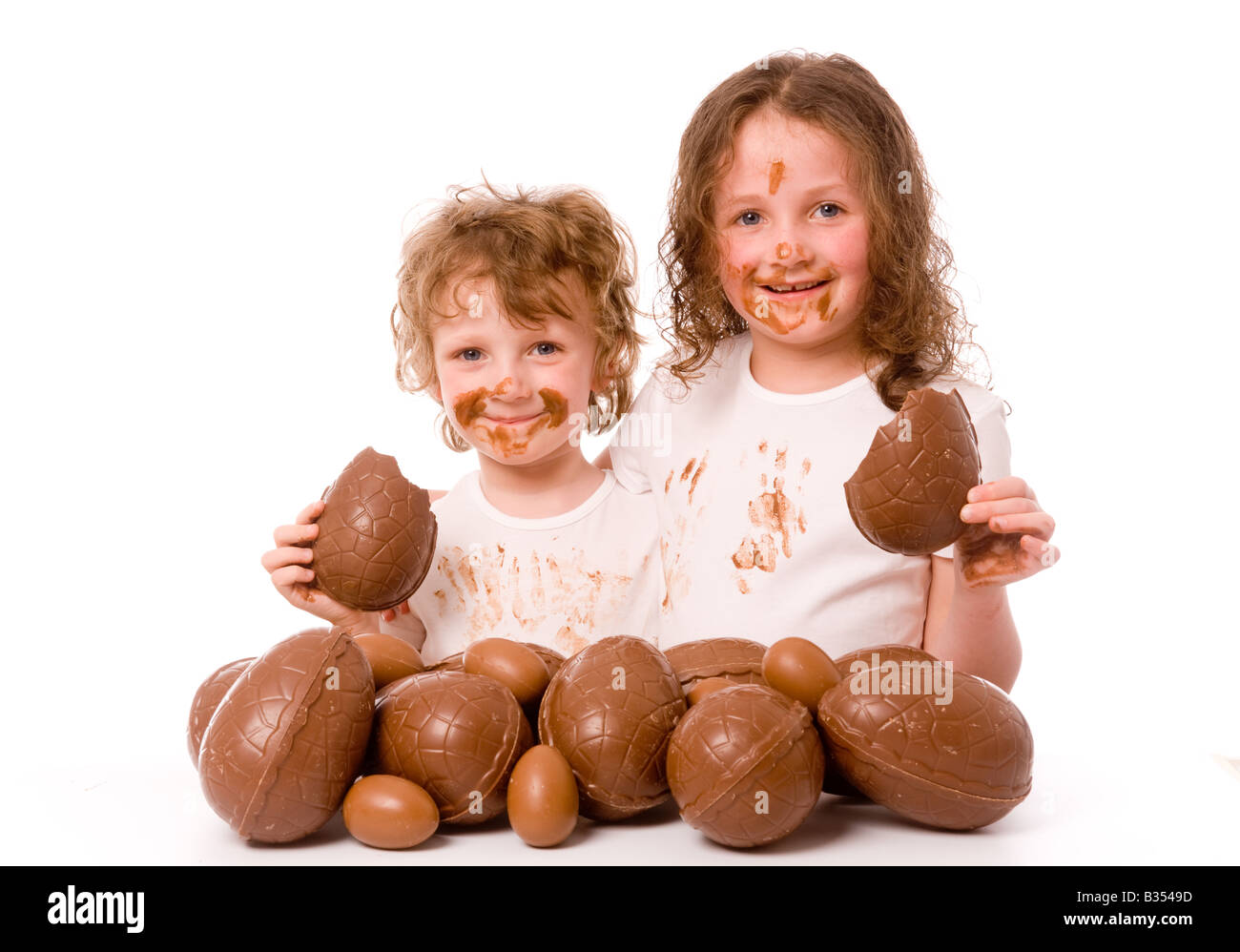 2 jeunes enfants avec du chocolat sur leur visage et leurs vêtements, chacun tenant un morceau d'oeuf de chocolat, avec beaucoup plus d'oeufs de Pâques au premier plan. Banque D'Images