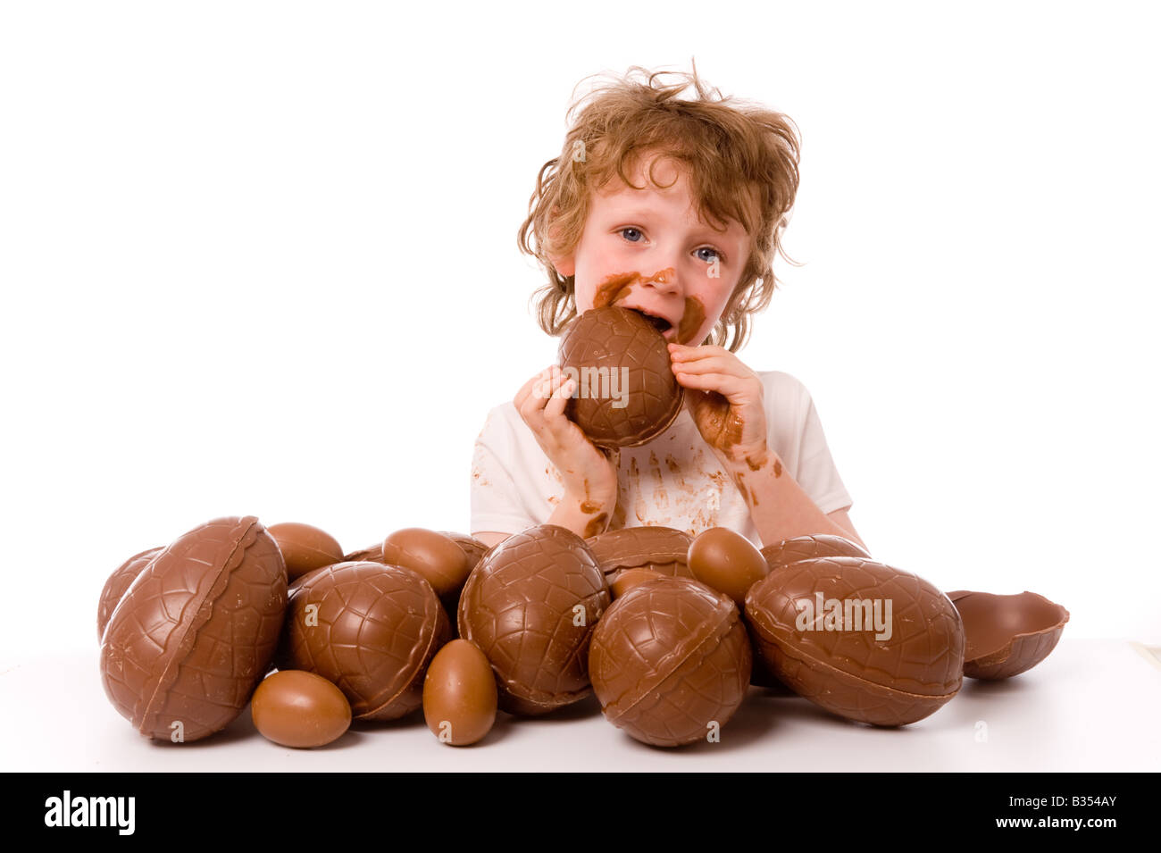 Jeune enfant aux cheveux courts désordonnés avec du chocolat sur les mains et le visage, mangeant un oeuf de Pâques avec plus d'oeufs non emballés au premier plan. Banque D'Images