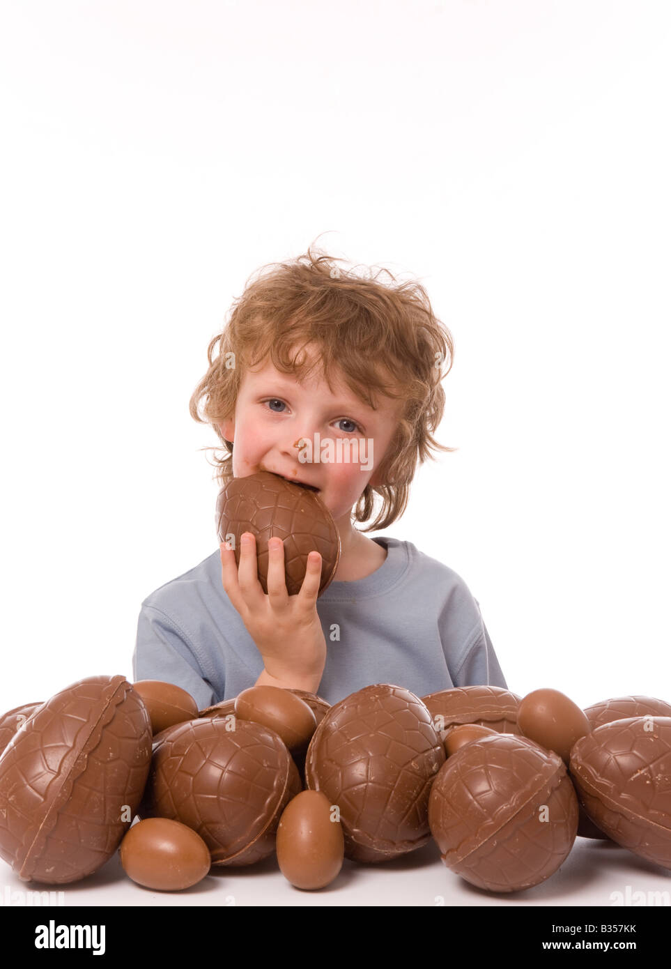Jeune enfant avec une grosse pile d'œufs de pâques, mangeant un œuf au chocolat sur le visage. Banque D'Images