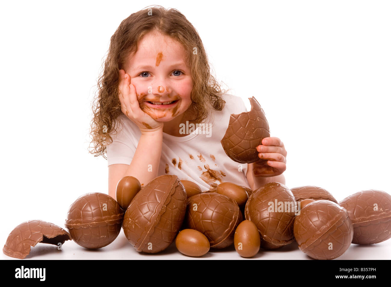 Jeune enfant souriant avec du chocolat sur son visage et t-shirt blanc avec beaucoup d'oeufs de Pâques au premier plan, vu sur un fond blanc. Banque D'Images