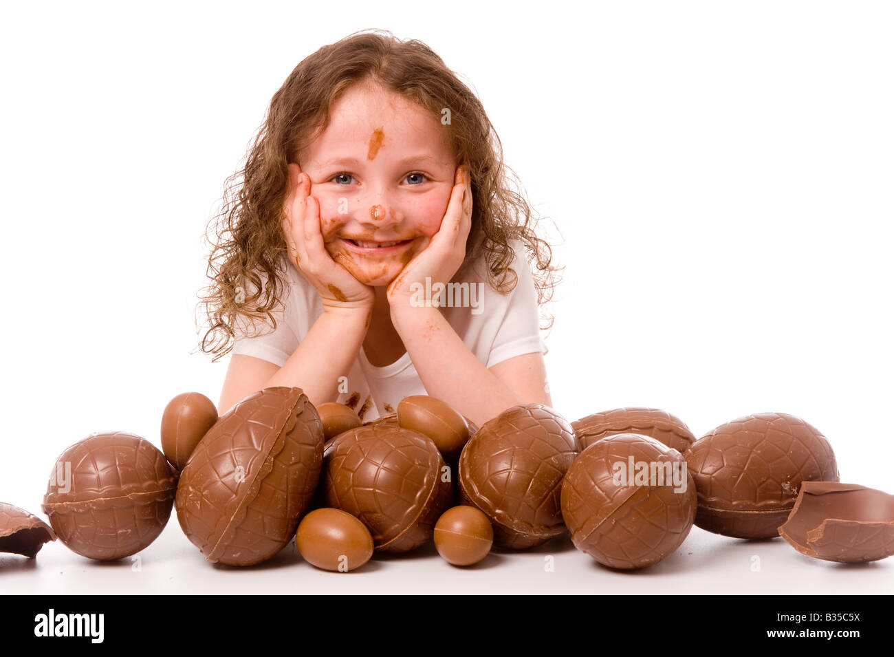 Jeune enfant souriant se reposant la tête dans ses mains enrobées de chocolat devant beaucoup d'oeufs de Pâques. Banque D'Images