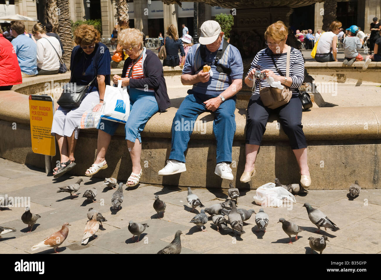 Espagne Barcelone quatre touristes matures s'asseoir sur la fontaine de Plaça Reial et manger avec les pigeons néoclassique à proximité square plaza Banque D'Images