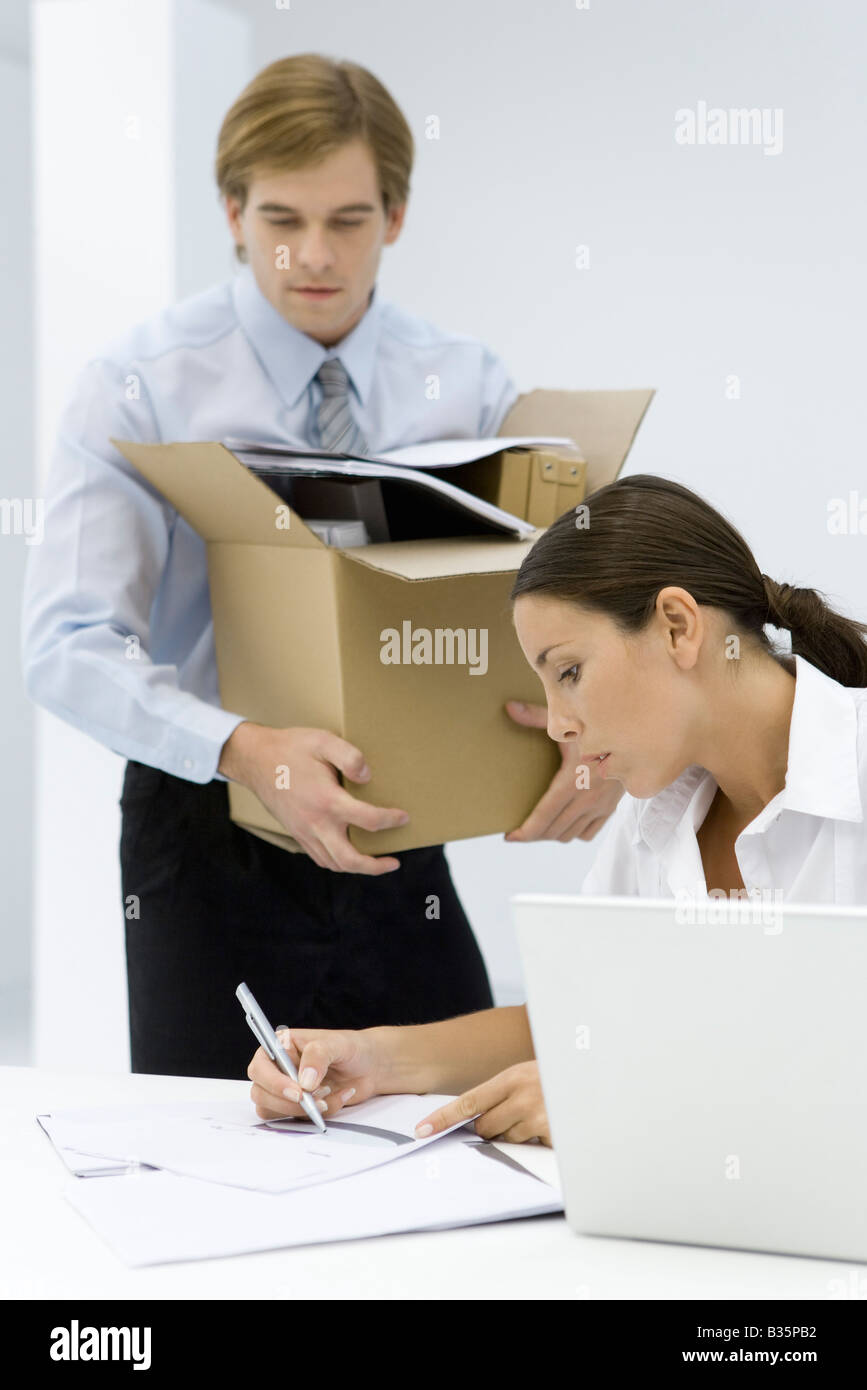 Femme écrit sur le document au bureau, man carrying cardboard box plein de fournitures de bureau Banque D'Images
