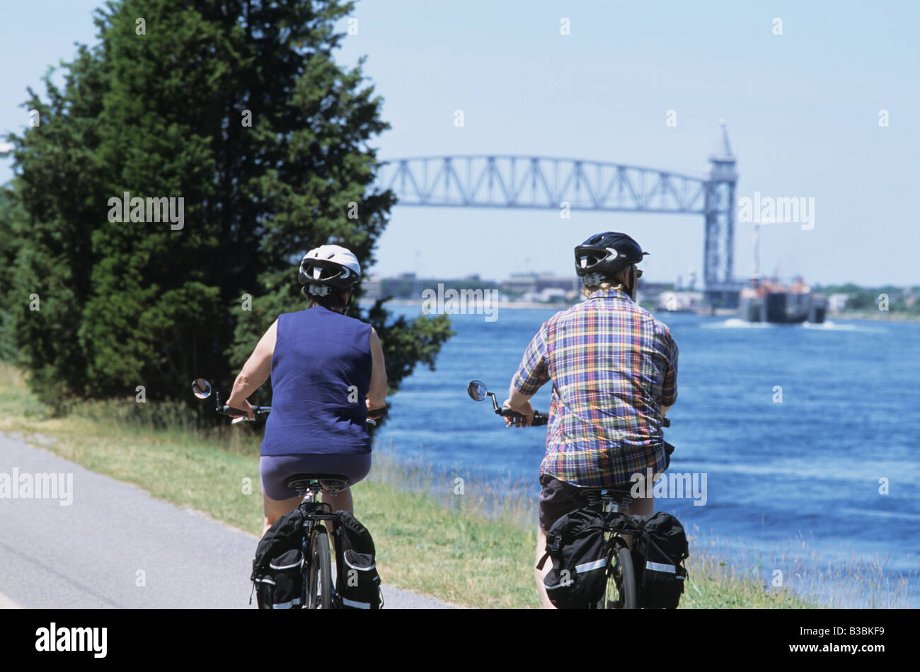 2 Deux personnes Homme et femme équitation sur des vélos avec casques bleus le long de la piste cyclable du Canal de Cape Cod avec pont de chemin de fer en arrière-plan Banque D'Images