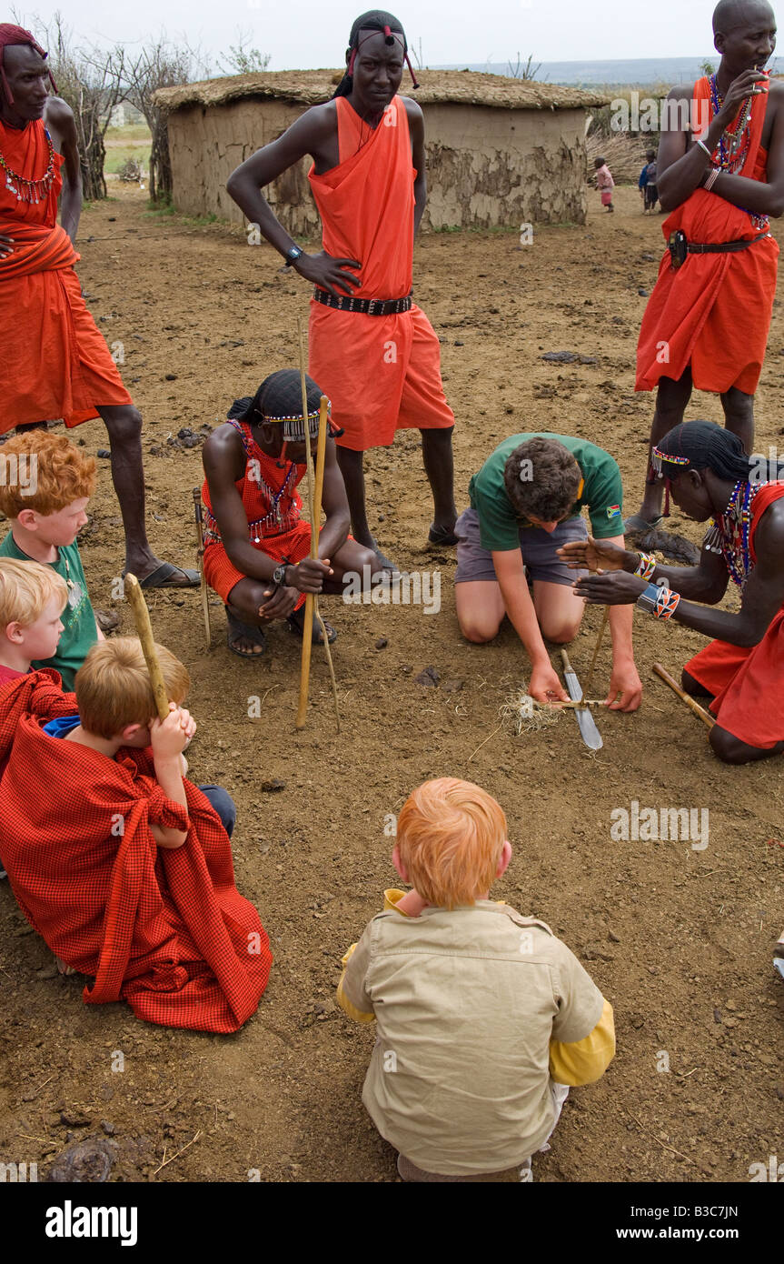 Kenya, Masai Mara National Reserve. Au cours d'une visite d'une manyatta Masaï enfants sur une famille safari regardez comme les guerriers démontrer faire feu en frottant des bâtons. Banque D'Images