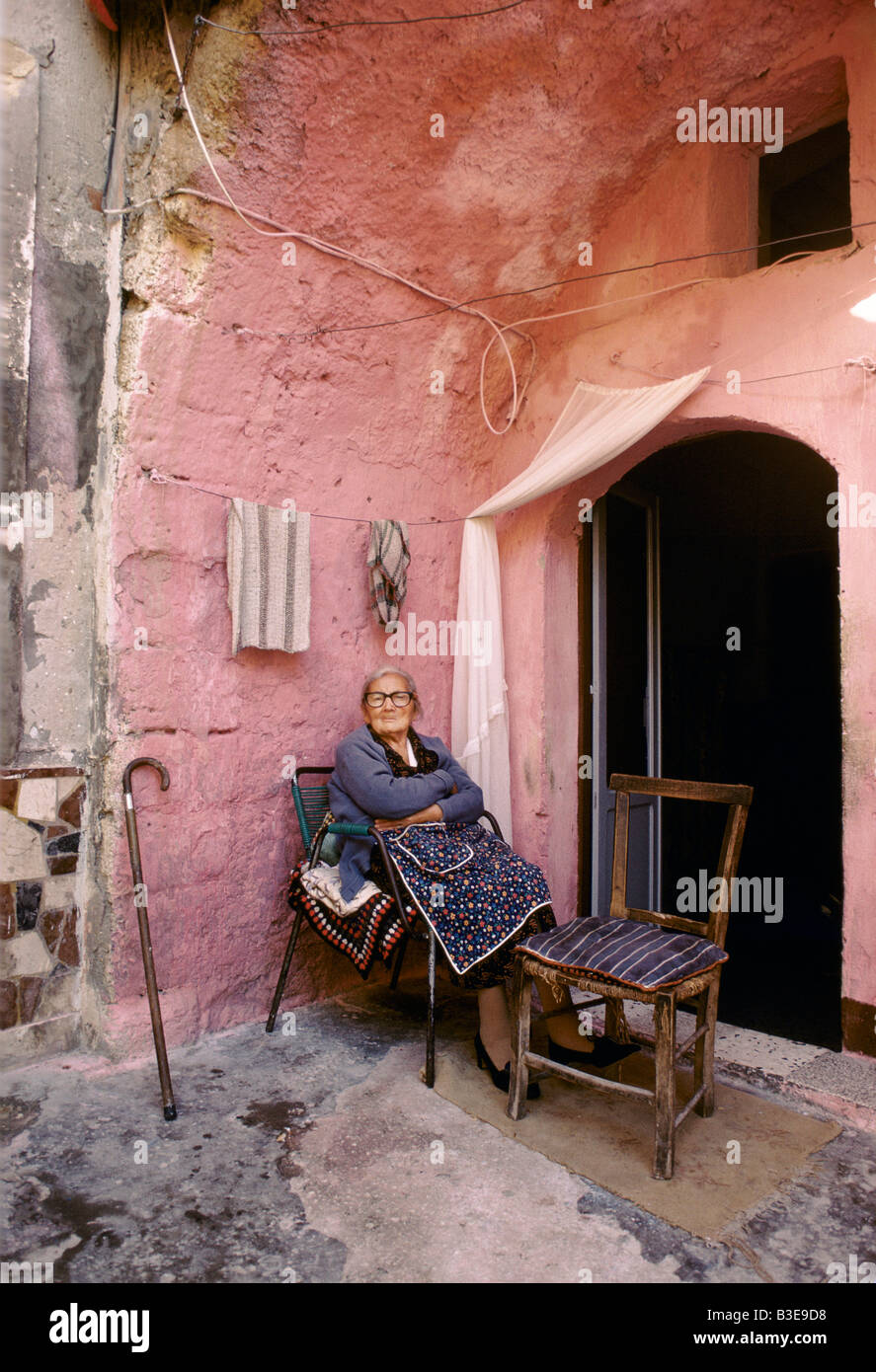 Une vieille femme EN TABLIER EST ASSIS SUR UNE CHAISE AVEC UNE CANNE APPUYÉE CONTRE LE MUR D'UNE ARCHE ROSE Banque D'Images