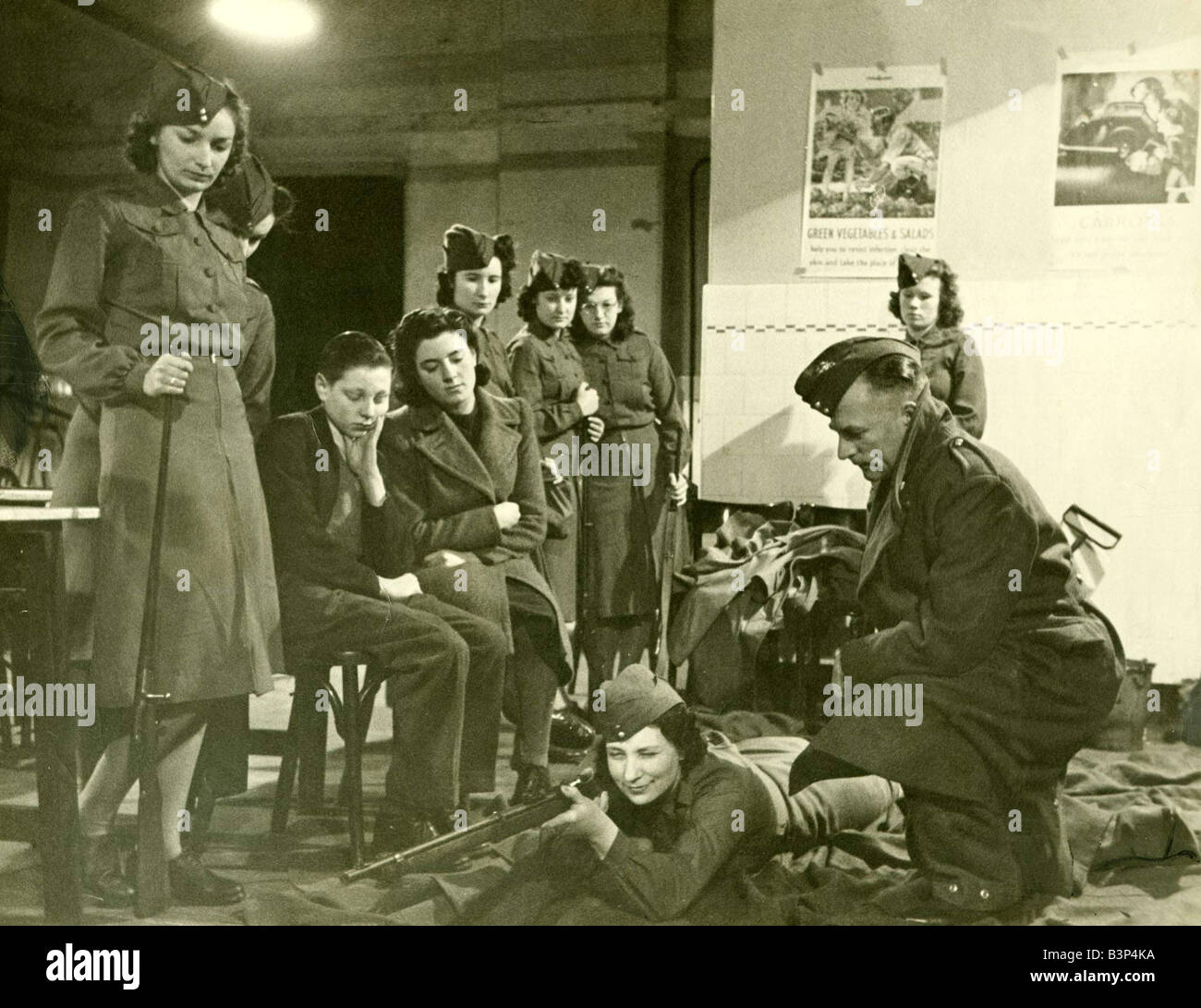 Mme Carson Olive en tenant la formation instructeur instruction de fusil tandis que deux de ses enfants, regardez sur l'armée britannique femme couchée sur le sol en vue d'une cible de mars 1942 Banque D'Images