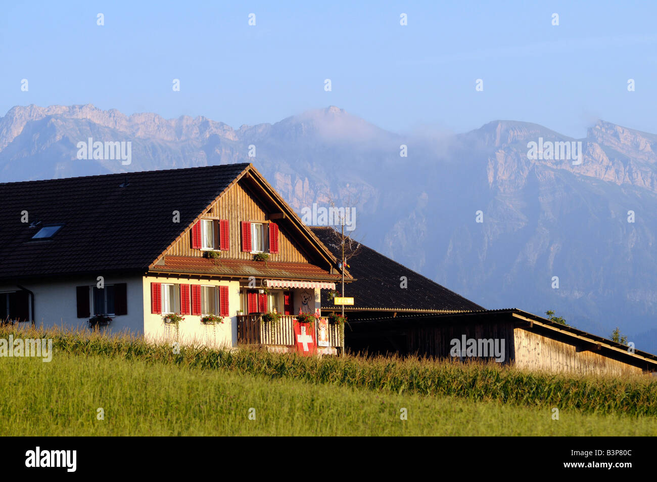 Une maison traditionnelle suisse typique dans un cadre rural, avec arbres, montagnes, et drapeau national ; en Suisse centrale Banque D'Images