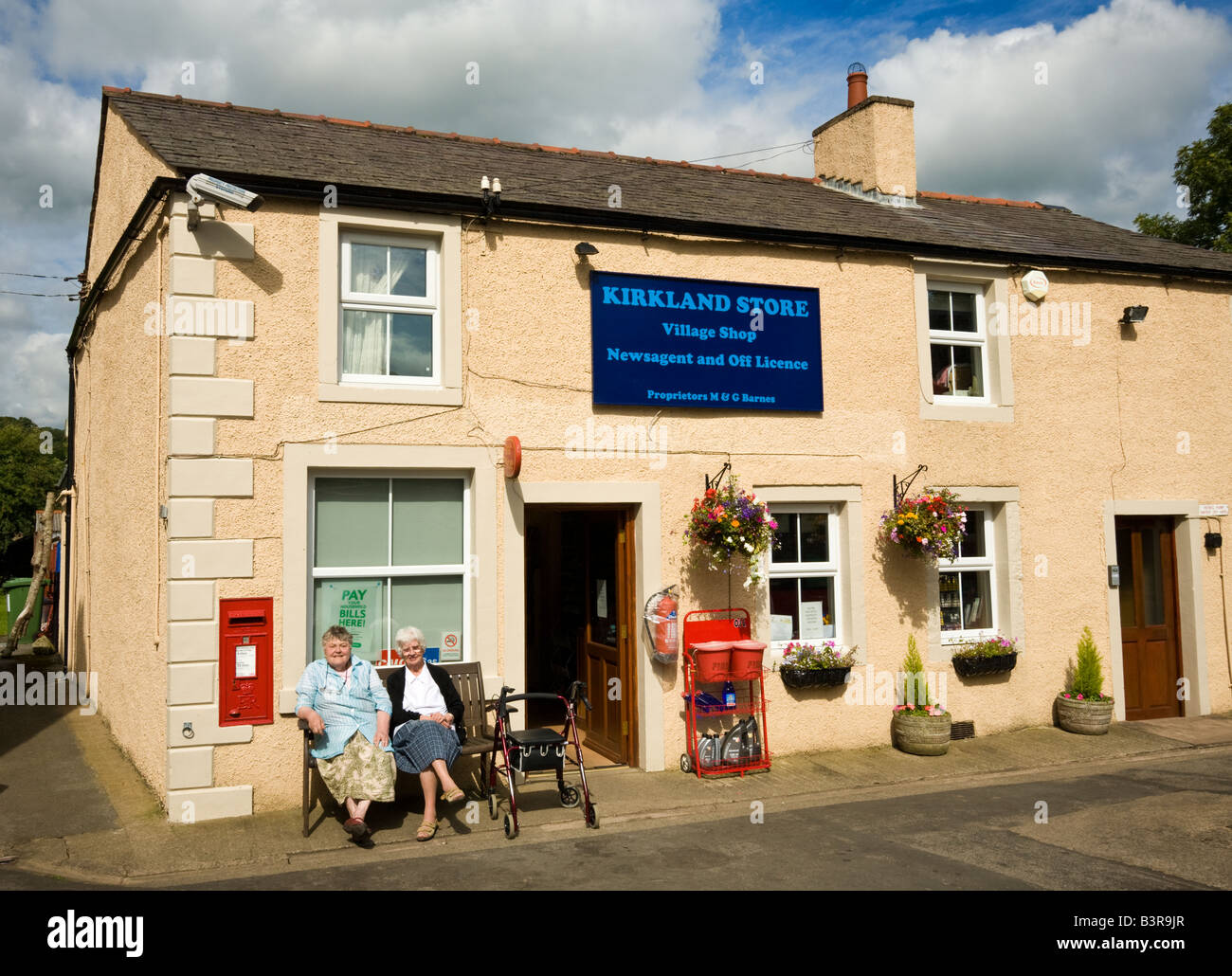 Magasin du coin, magasin de village rural et bureau de poste à Caldbeck, Cumbria, Lake District, Angleterre, Royaume-Uni avec deux retraités profitant du soleil à l'extérieur Banque D'Images