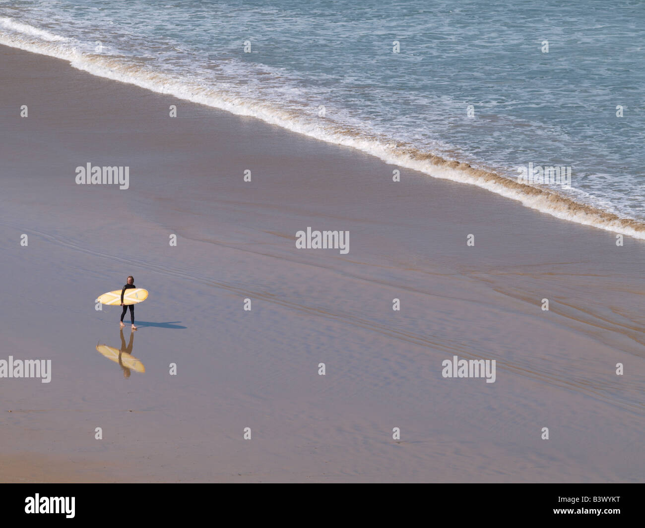 Surfeur homme marchant le long de la plage avec sa planche de surf sous le bras, reflète clairement dans le sable de l'eau. Newquay, Cornwall Banque D'Images