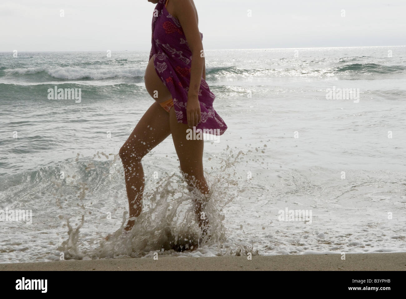 Femme enceinte marchant le long de la plage éclaboussant de l'eau en  portant un chemisier sur un maillot de bain. Canet Plage nr Perpignan Sud  de la France 2008 Photo Stock -