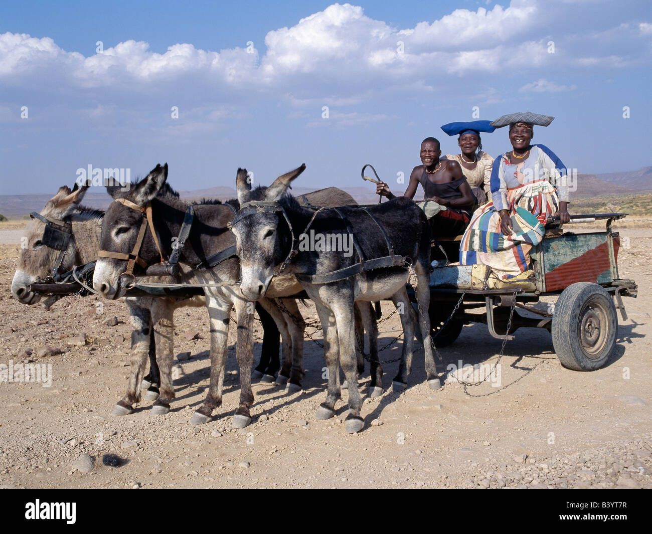 La Namibie, Damaraland, Palmwag. Un homme et deux femmes Herero ride home dans une charrette à âne. L'élaboration des robes et de styles uniques hat femmes Herero remonte au 19ème siècle les missionnaires allemands qui se sont opposés à ce qu'ils considéraient comme une forme de modestie parmi la tribu de robe. Ils ont insisté pour que les femmes Herero peu pratique d'adopter leur propre style de robes de l'époque victorienne. Au fil des ans, robes Herero colorés ont évolué aussi énorme crinolines porté sur cinq jupons. Banque D'Images