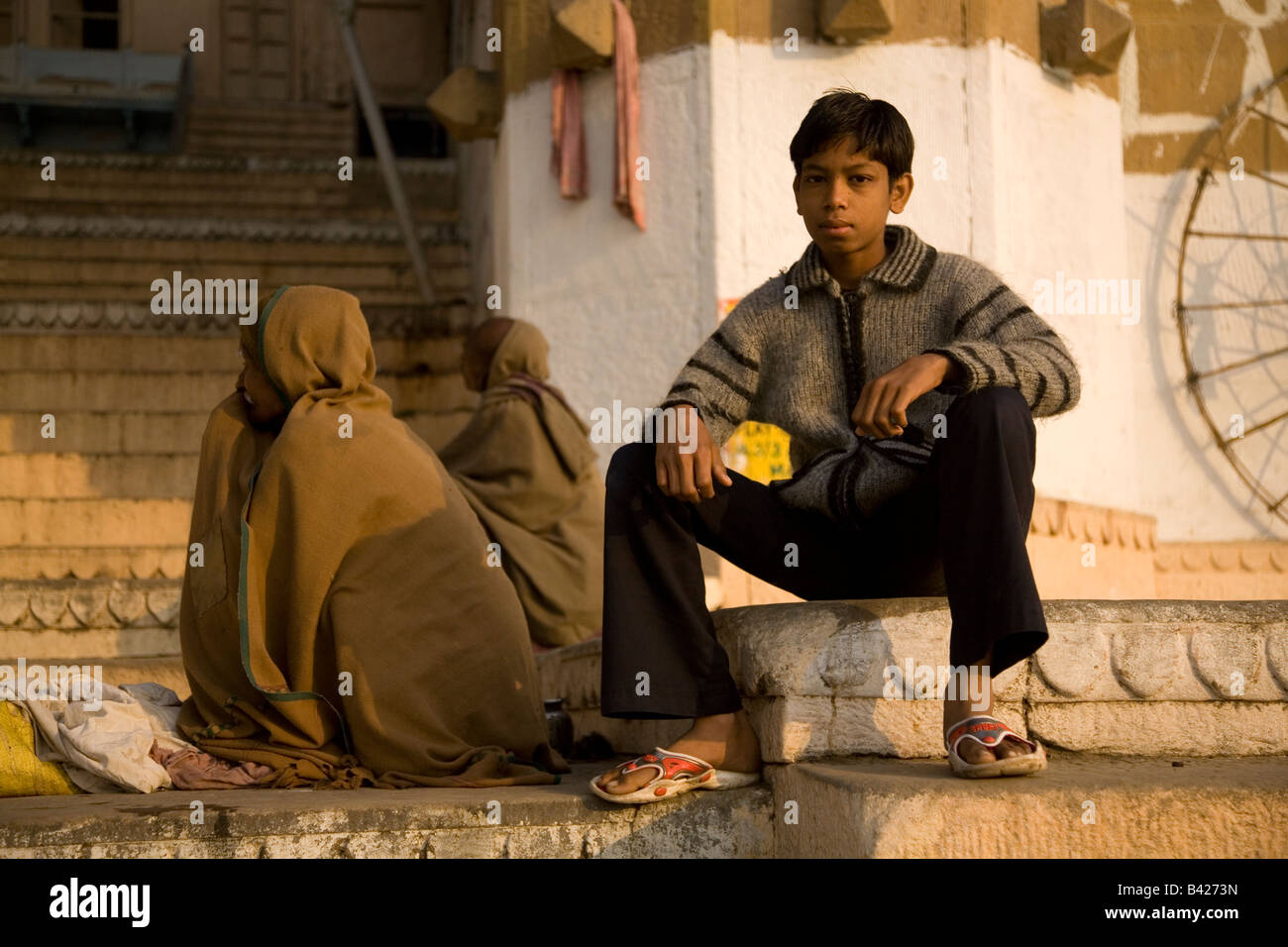 Un garçon est assis sur l'un des Ghats dans la ville de Varanasi, en Inde. Deux hommes wearning capes s'asseoir derrière lui. Banque D'Images