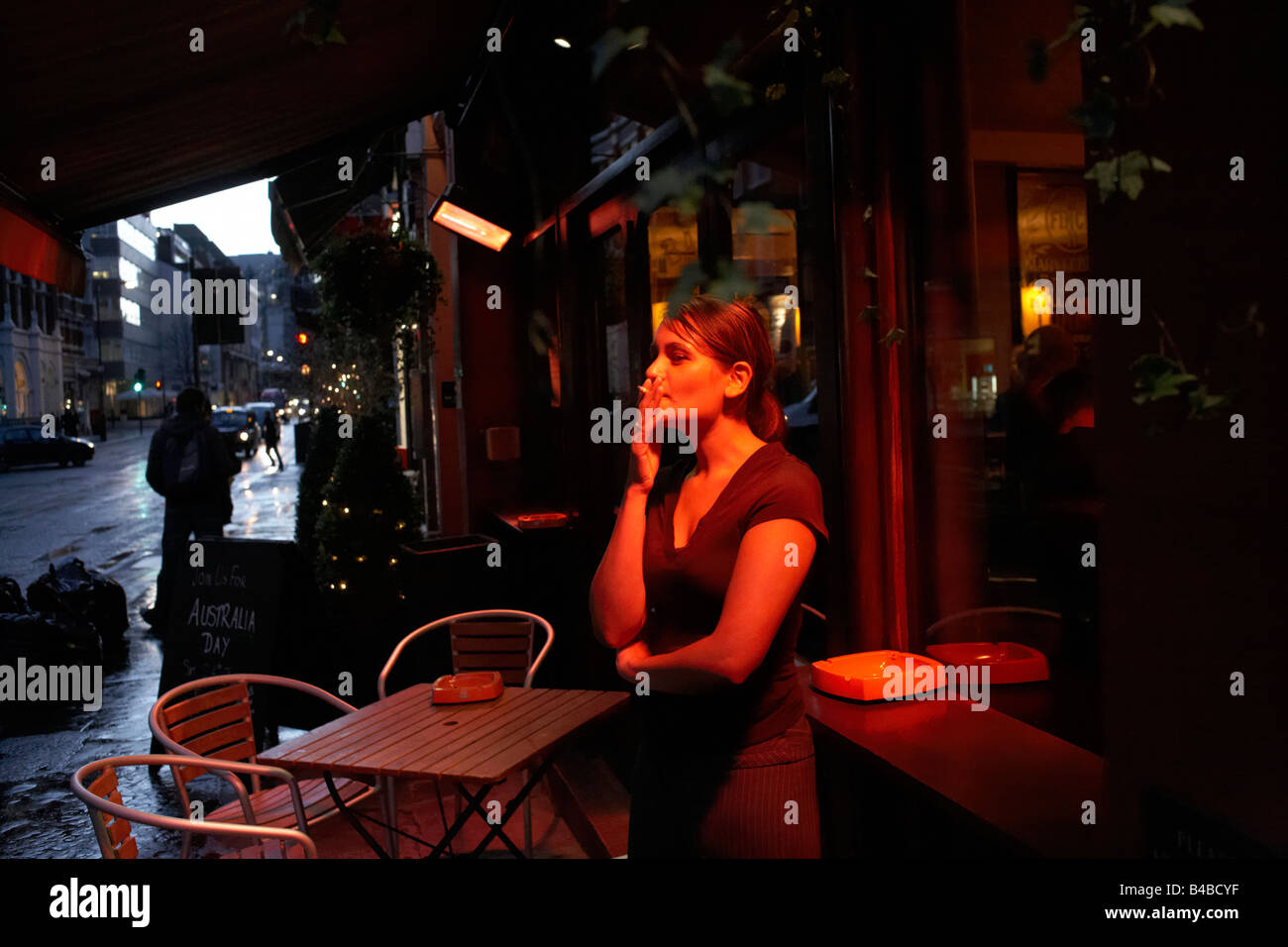 Sur une nuit pluvieuse, une jeune femme fume sous un abri chauffé zone pour fumeurs à l'extérieur d'un bar à Londres Banque D'Images