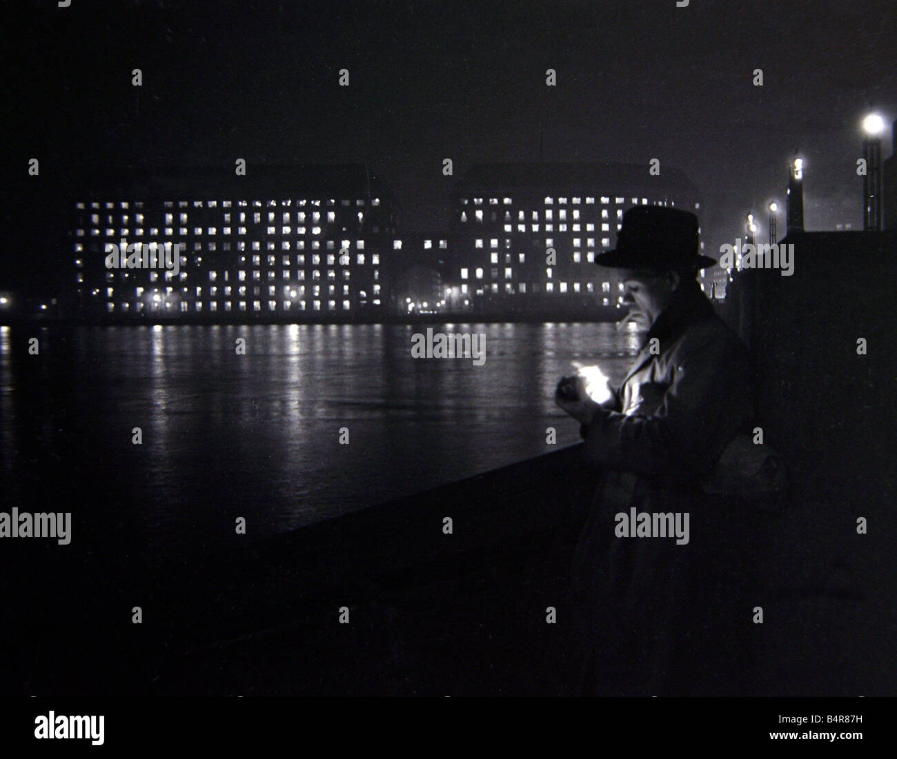 Un homme allume une cigarette sur le quai de la Tamise à Londres vers 1955 personnes Secret Homme Bâtiments Cigarette nuit 1950 Detective Moody Spy Banque D'Images