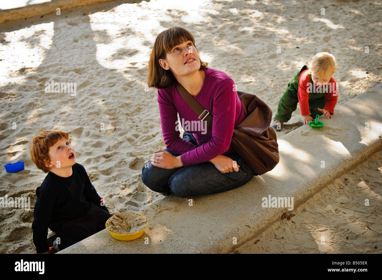 Mère assise avec ses deux jeunes fils qui jouent dans un bac à sable. Banque D'Images