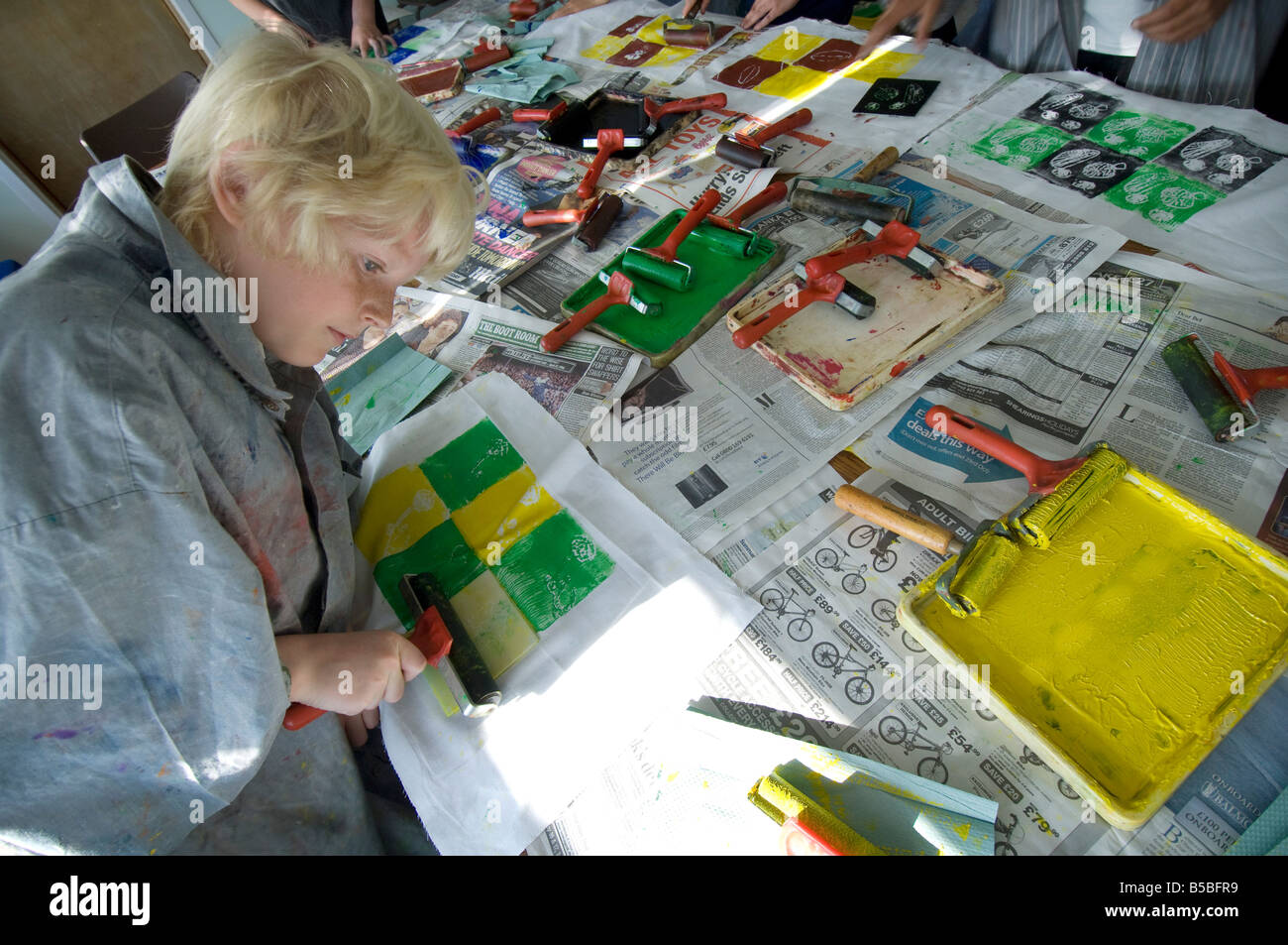 Un écolier aux cheveux juste dans une classe d'art à l'aide d'un rouleau à l'encre verte et jaune des carrés. Banque D'Images