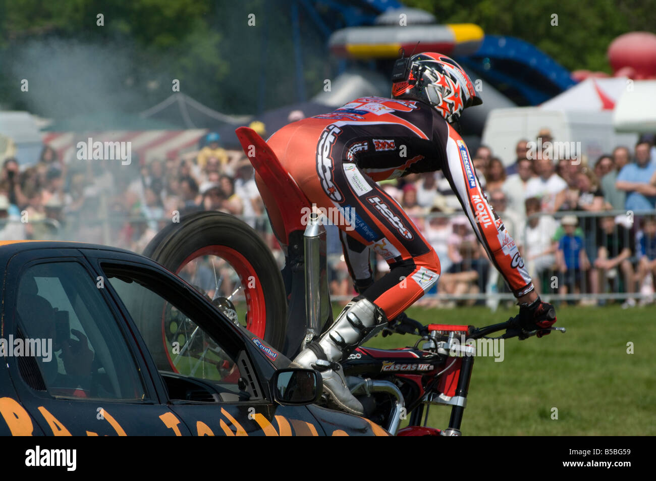 Stunt moto Rider équitation sa moto produisant de la fumée des cascades de la scène sa roue arrière sur une voiture de rallye Bonnet Cowpie Betchworth Surrey Banque D'Images