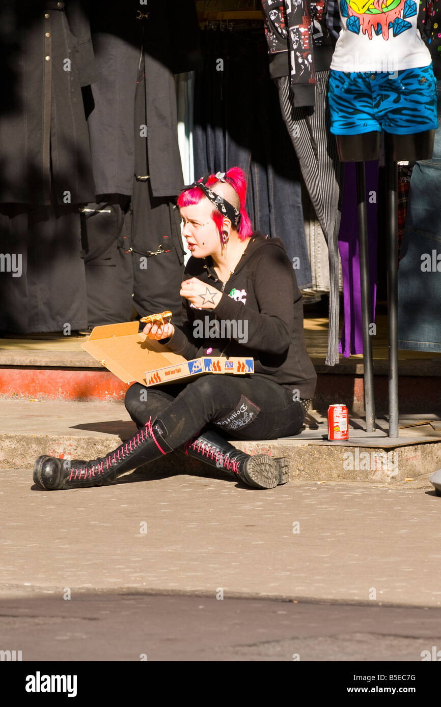 London Camden Lock Market jolie fille punk cheveux rose queue partie bottes tête rasée percés bouche mange pizza by boutique Banque D'Images
