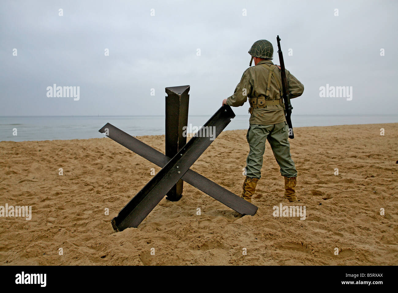 Un acteur habillé comme un soldat américain D-Day sur la plage Utah Normandie France debout à côté d'un réservoir 'Hedgehog' piège. Banque D'Images