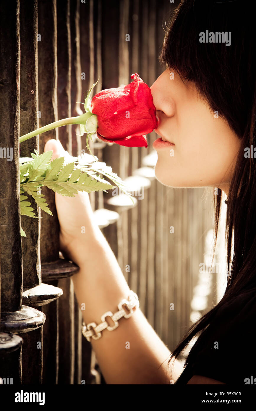 Jeune femme anonyme qui sent une rose rouge derrière clôture métallique Banque D'Images