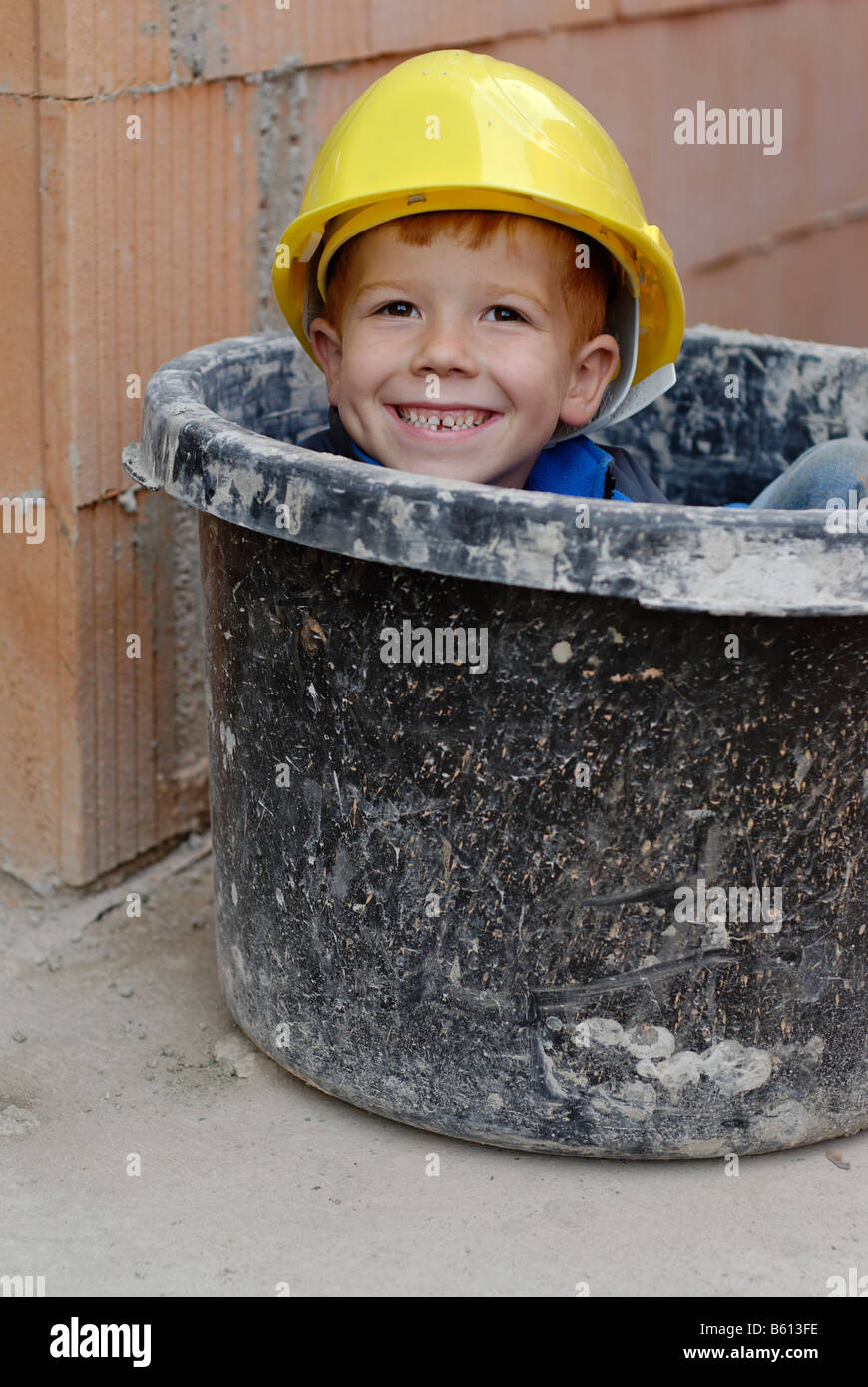 Little smiling boy portant un casque jaune assis dans un mortier de plâtre seau à la maison construction site Banque D'Images