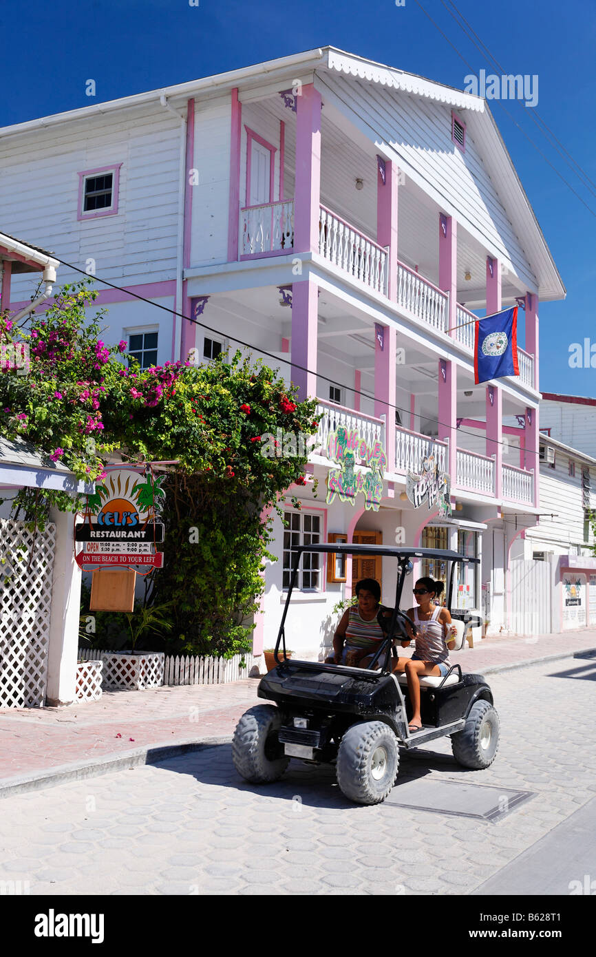 Deux femmes dans une voiturette de golf qui passent une maison en bois peint de couleurs des Caraïbes, San Pedro, Ambergris Cay Island, Belize, 100 Banque D'Images