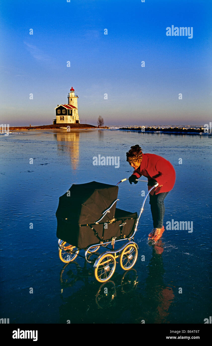 Les Pays-Bas, Marken. Patinage mère avec bébé buggy sur lac gelé appelé IJsselmeer. Contexte : le phare. Banque D'Images