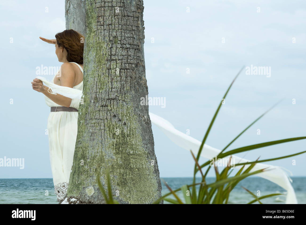 Femme debout à côté de palmier, ombrage les yeux avec la main, à la recherche en mer, d'écharpe circulant dans Breeze Banque D'Images