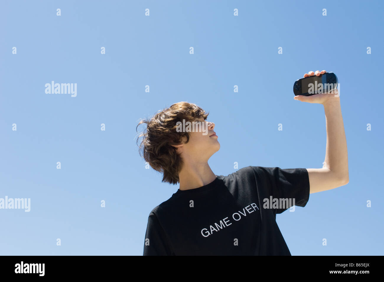 Teen boy holding up jeu vidéo de poche, portant des tee-shirt imprimé avec les mots 'game over' Banque D'Images