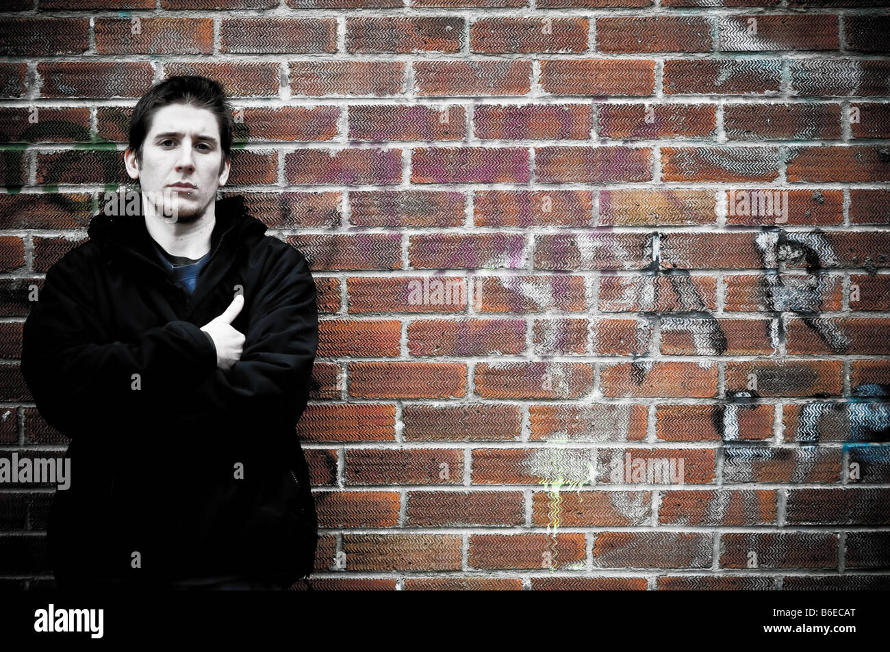 Jeune homme s'appuyant sur un mur de briques couverts de graffitis, les bras croisés, à la UK agressif Banque D'Images