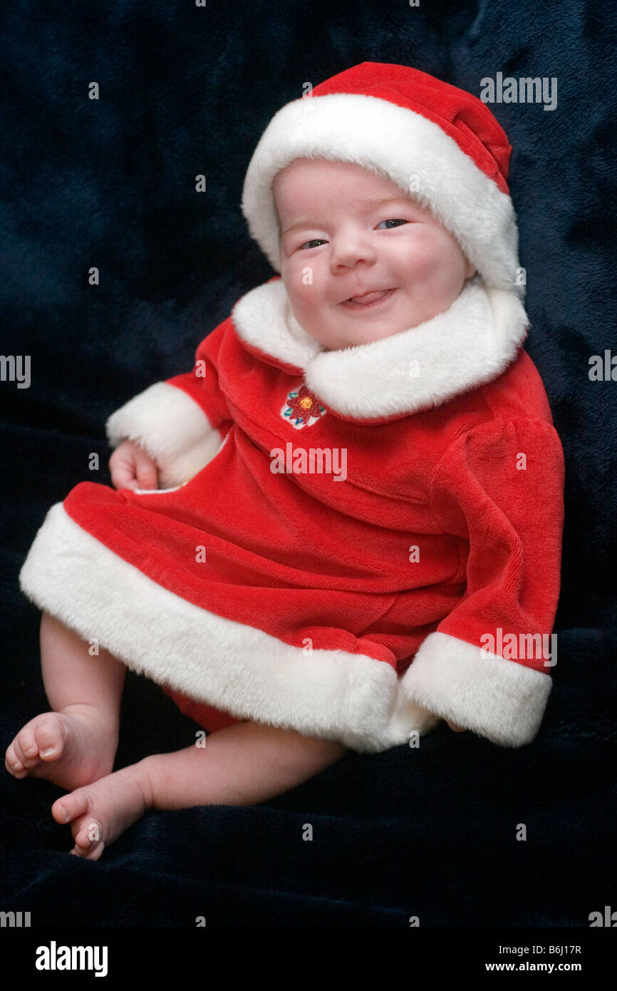 Trois mois baby girl smiling, habillé dans une robe et Santa hat pour Noël sur un fond sombre Banque D'Images