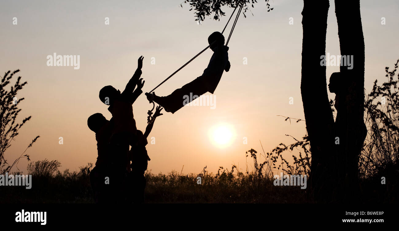 Silhouette d'enfants indiens jouant sur une balançoire fait maison à la campagne au coucher du soleil. L'Andhra Pradesh, Inde Banque D'Images