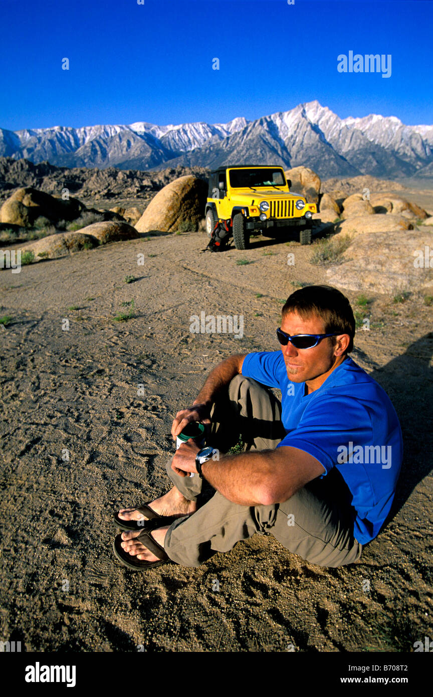 L'homme avec des lunettes de soleil assis dans le désert, à côté de sa jeep en face de la montagne. Banque D'Images