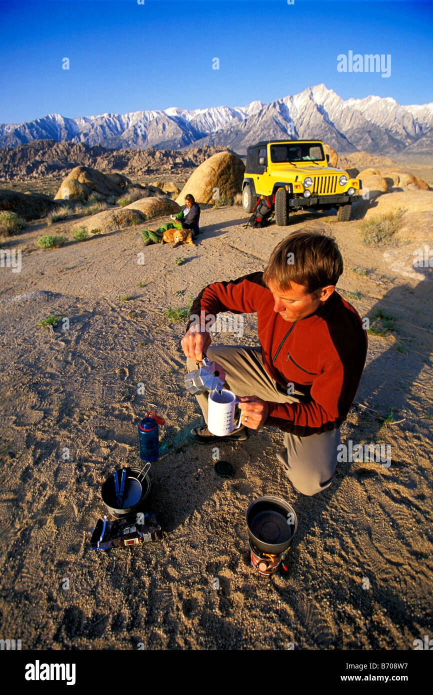 L'homme faire le café sur un réchaud dans le désert avec une femme, jeep et les montagnes en arrière-plan. Banque D'Images