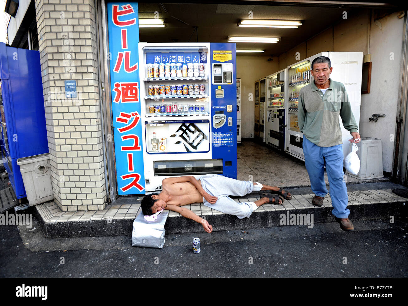 Un sans-abri se trouve endormi par distributeurs automatiques de bière bon marché et de vin de riz saké tandis qu'un autre homme marche passé à Osaka au Japon Banque D'Images
