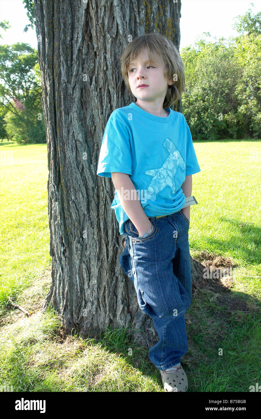 Un garçon de six ans avec les mains dans les poches se situe à côté de l'arbre, Winnipeg, Canada Banque D'Images