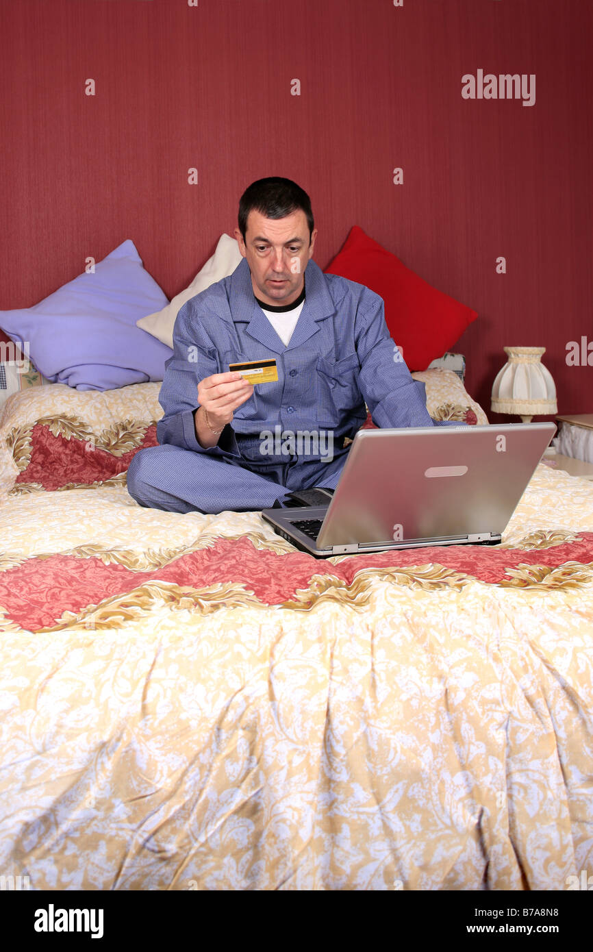 Homme assis sur son lit wearing pajamas holding d'une carte de crédit à l'aide d'un ordinateur portable pour faire un achat en ligne tôt le matin Banque D'Images