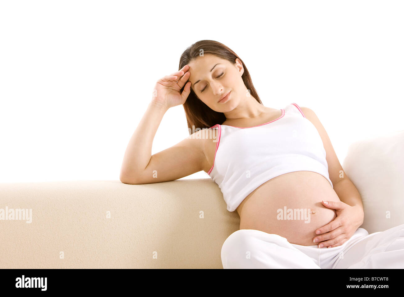 Jeune femme enceinte assise sur canapé les yeux fermés, une main sur l'abdomen Banque D'Images