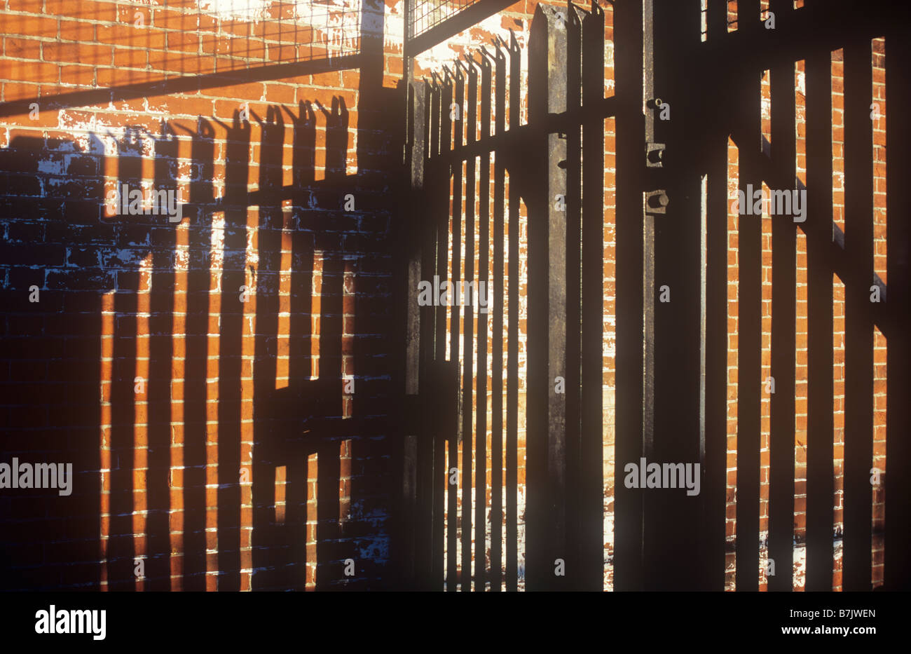Black metal haute sécurité surmontées de pointes inclinées et treillis métallique d'ombres profondes sur mur brique orange en lumière chaude Banque D'Images