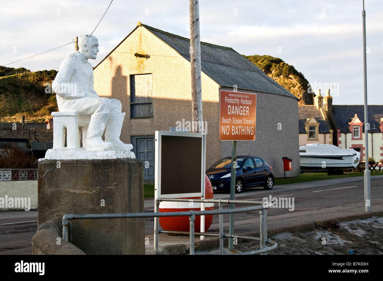 Statue d'un homme assis sur un banc à côté d'un panneau d'avertissement rouge à Findochty, en Ecosse Banque D'Images Statue d'un homme assis sur un banc à côté d'un panneau d'avertissement rouge à Findochty, en Ecosse Banque D'Images