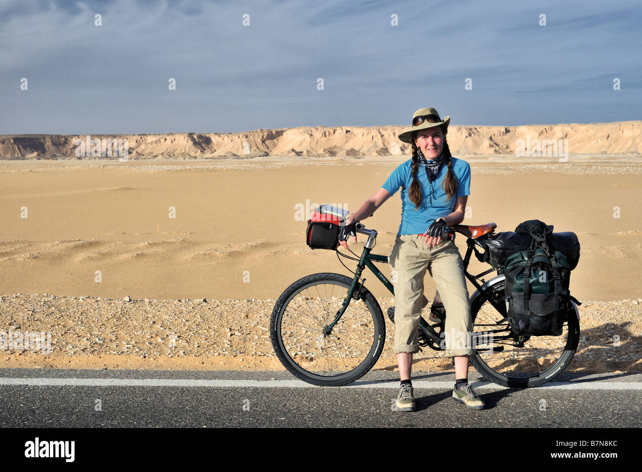 Western touring cyclist girl leaning contre vélo chargé dans désert occidental oasis road, dunes de sable/collines en arrière-plan Banque D'Images